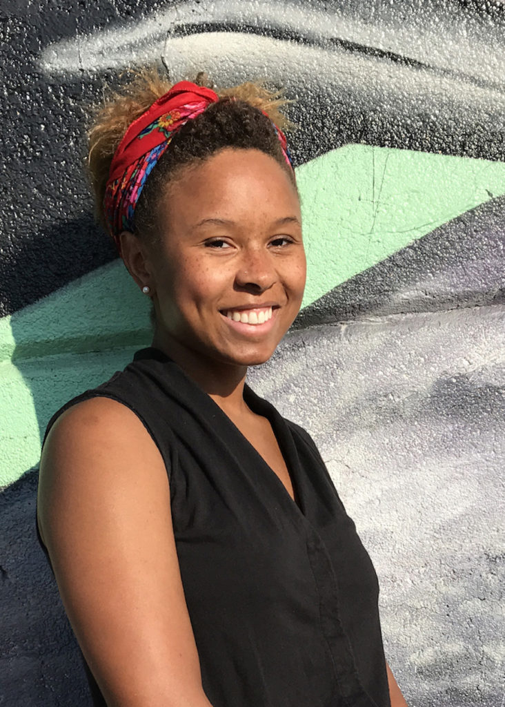 Marisa Williamson; a black woman with her hair pulled back by a red bandana; wearing a black tank top; standing in front of a grey and pale green mural