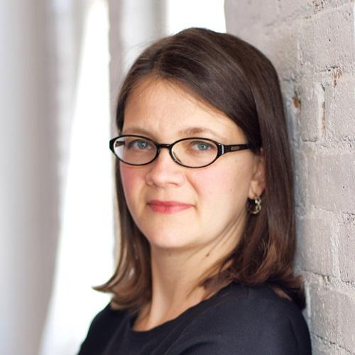 Headshot of Sarah Kirk Hanley, white woman with shoulder length brown hair and thin black glasses, leaning against a white brick wall