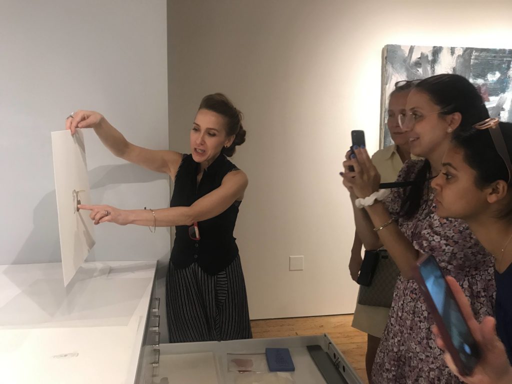 A woman holds up an art print and gestures to the middle of it while 3 other women look on and take photos.
