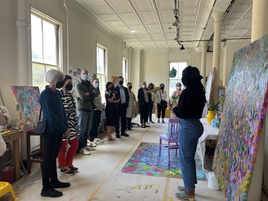 Inside an artist's studio, about 12 people face the artist and a large painting.