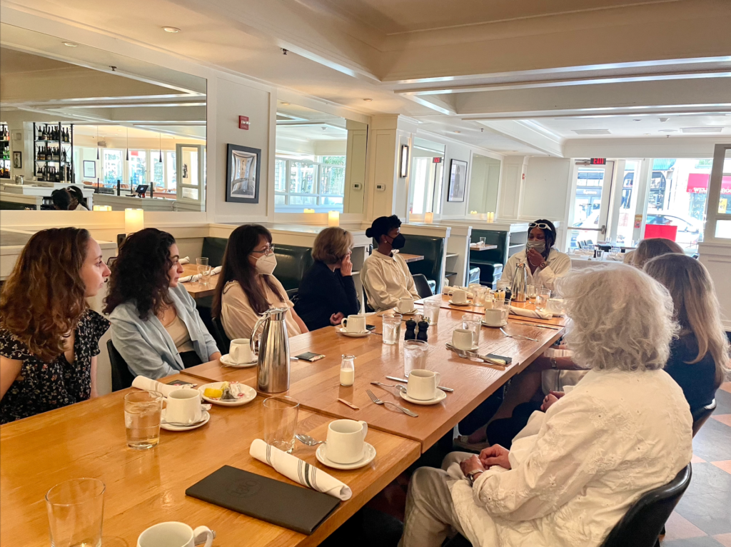 A group of women sit at a long table, listening to a speaker.