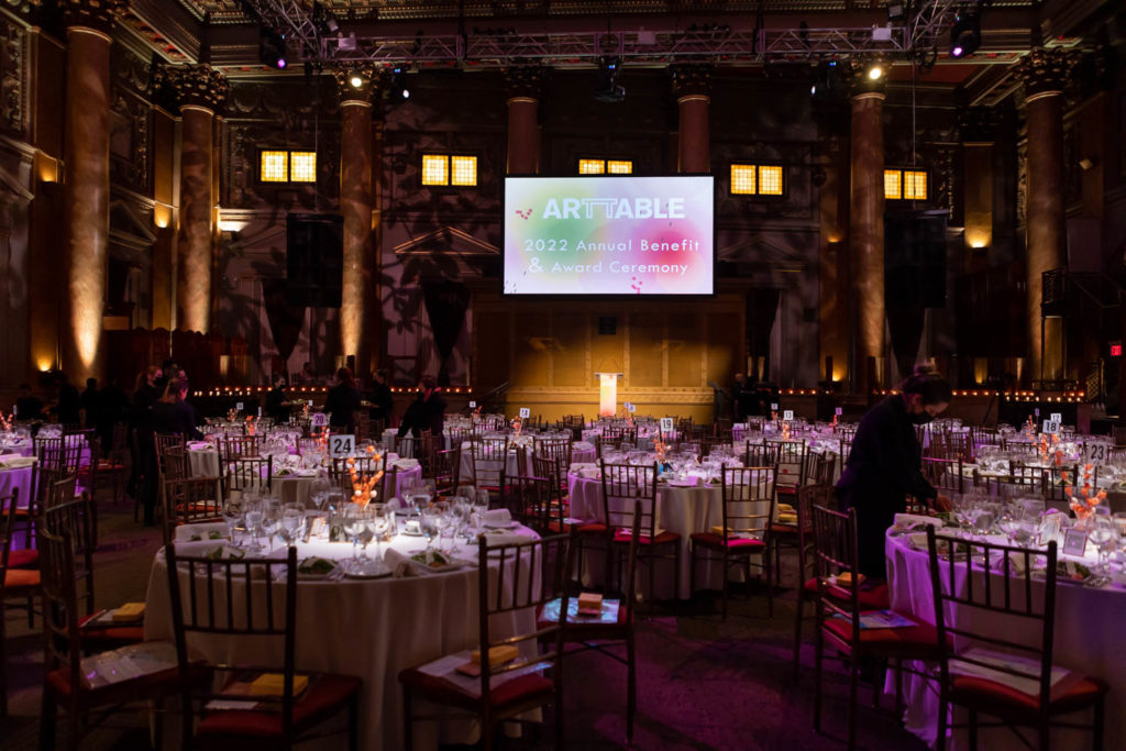 Interior at ArtTable's Annual Benefit, showing several large round tables with chairs, set for a luncheon. A large projector screen with ArtTable's logo is high on the back wall.