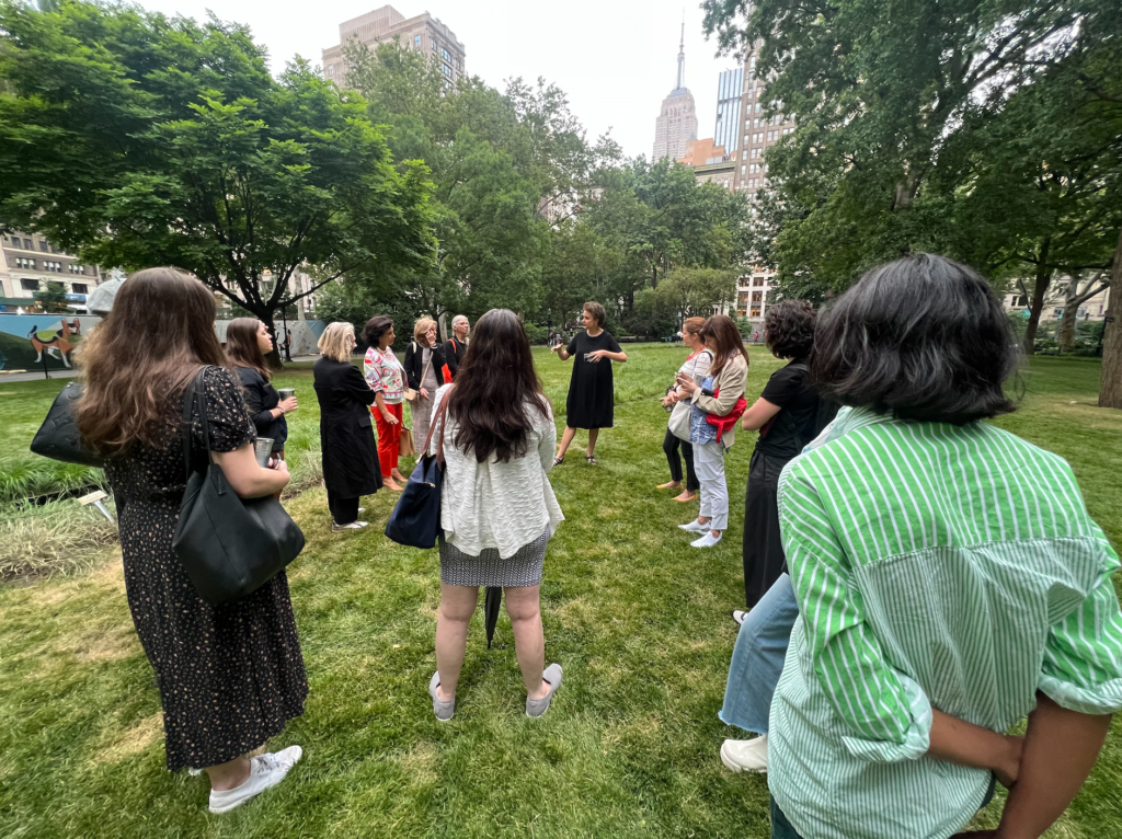 A view of a group from the back, facing a woman who is speaking. They are in Madison Square Park. The Empire State Building is visible in the background.