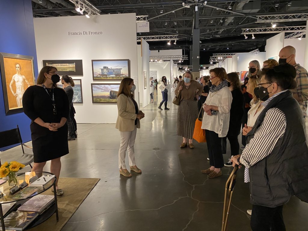 A group of people face a single woman who is assumedly speaking to them about the artwork on view in her art fair booth.