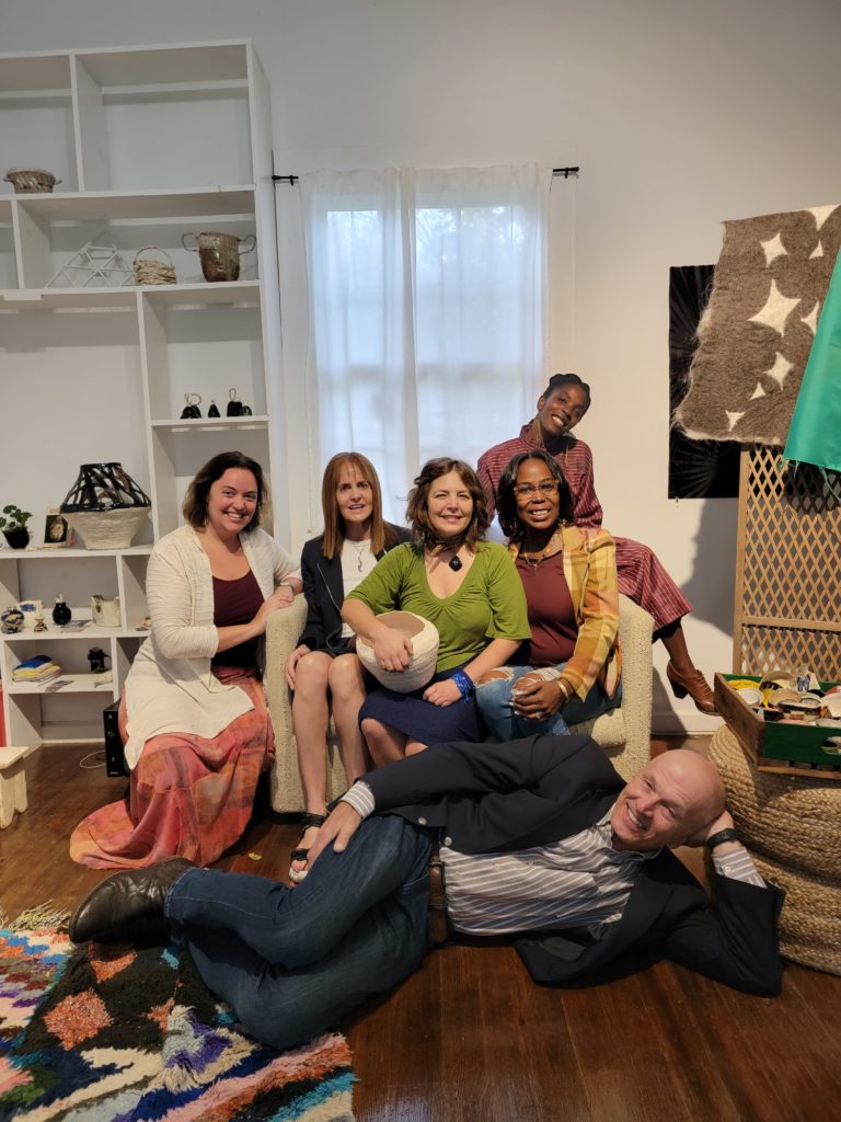 A group of 5 women sit on a loveseat smiling at the camera. A man lays sideways in front of them on the floor, leaning on his elbow. 