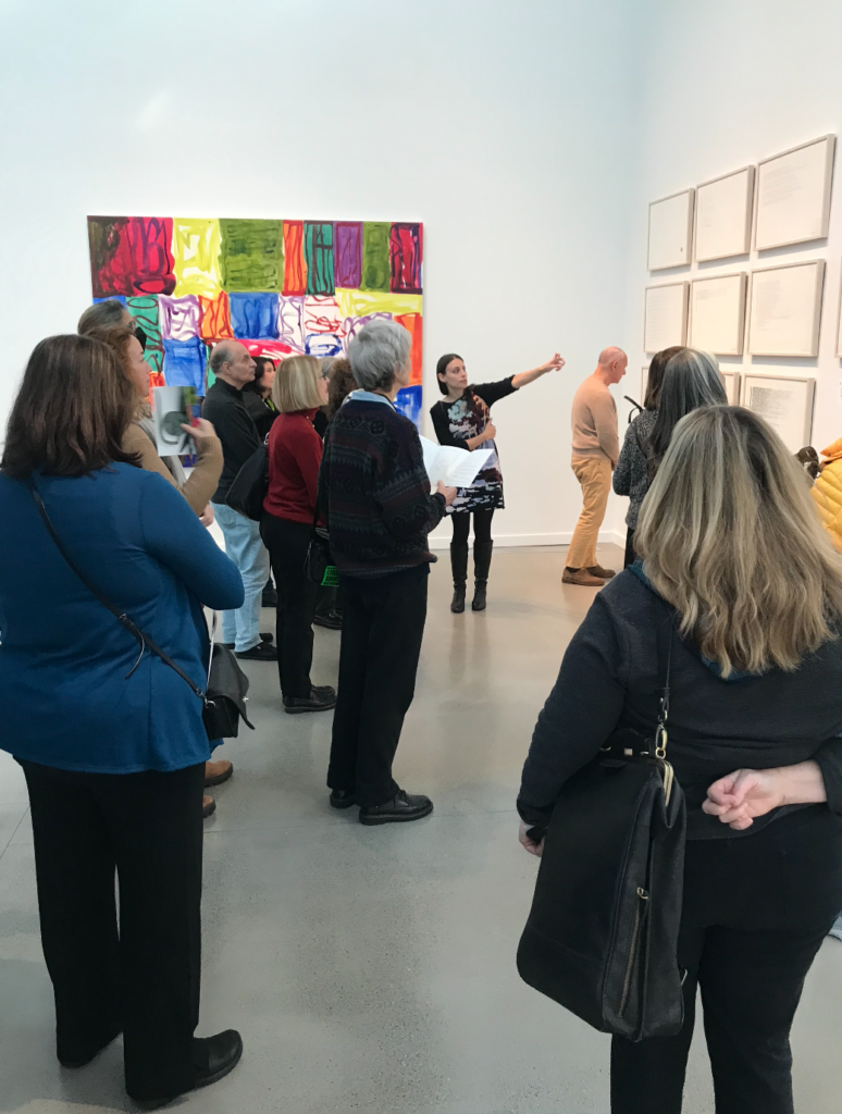 A woman gestures to some drawings on a wall while a group looks on and listens.