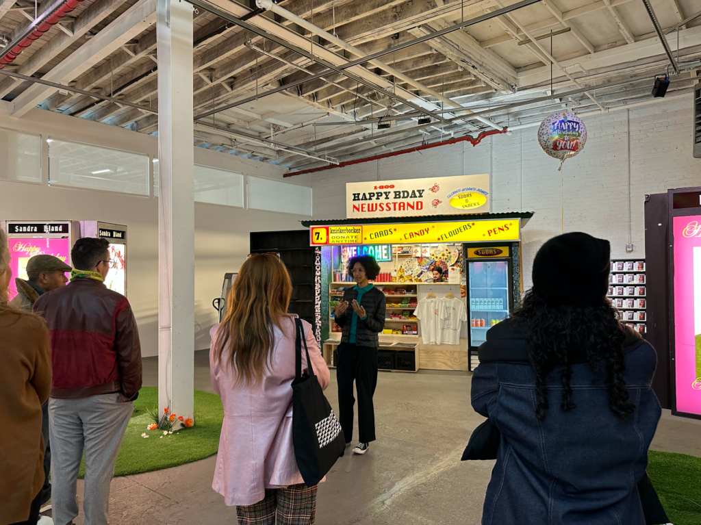 A group of 4-5 people stand with their backs to the camera, facing Klaudia Ofwona Draber who is speaking about the exhibition. Behind her is an art installation of a typical New York bodega.
