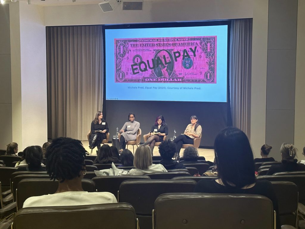 4 panelists seated on a stage with a large "Equal Pay" image on the screen behind them
