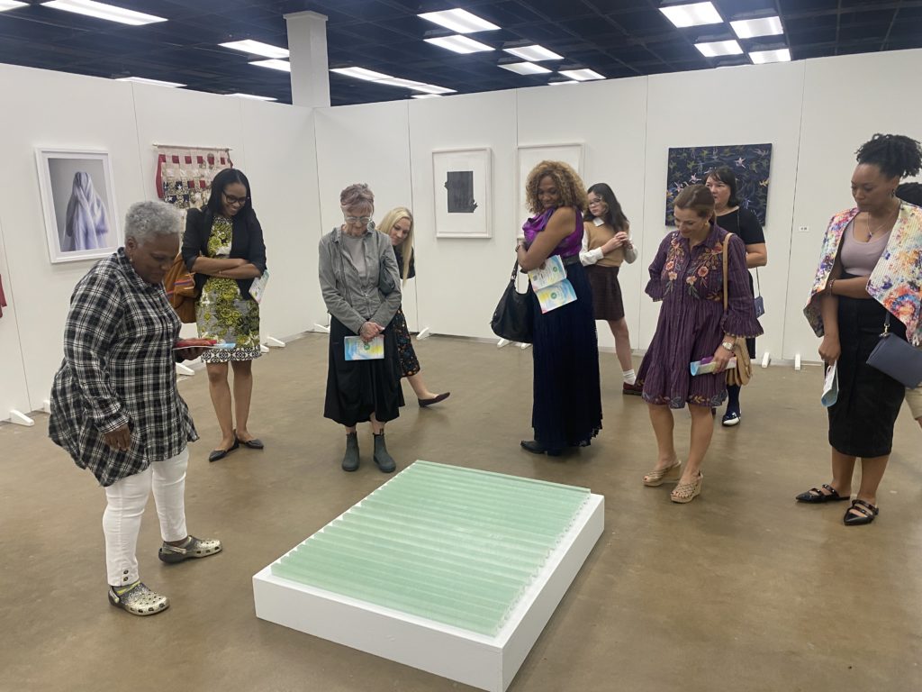 A group of women look down at a piece of artwork, a glass-looking sculpture.