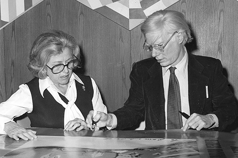 Black-and-white image of Marcia Simon Weisman, a white woman wearing black glasses, a black vest, and a white blouse, sits at a table with Andy Warhol, a man wearing clear glasses, a white shirt, and dark suit and tie. They are sitting in a wood-paneled room and looking document laid on the table.