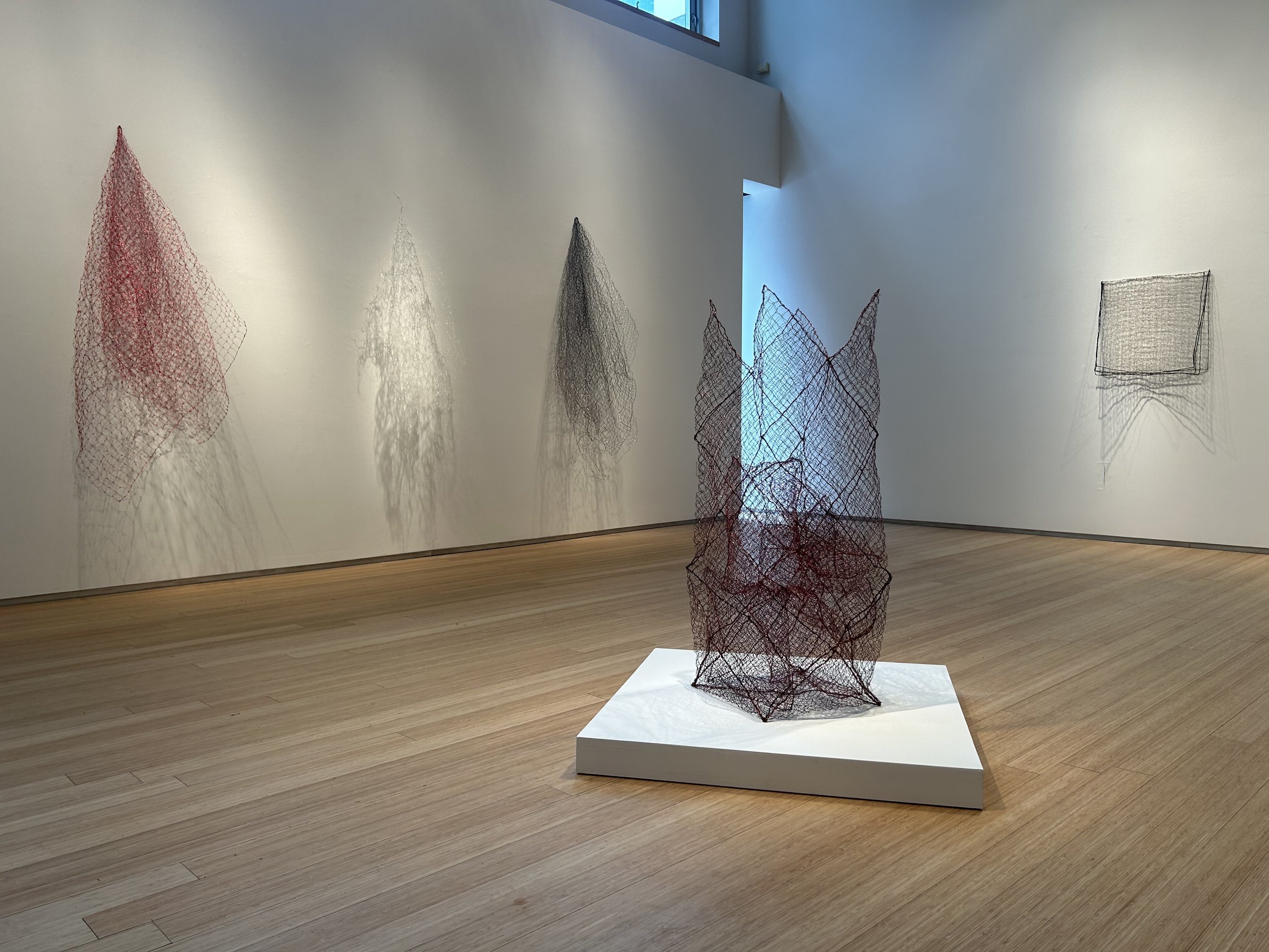 Installation view of Nancy Koenigsberg: Light and Shadow at Nancy Hoffman Gallery. Wire sculptures by Nancy Koenigsberg decorate a large room with white walls and a wood floor; four sculptures hang on the walls and a fifth sculpture stands in the middle of the room.