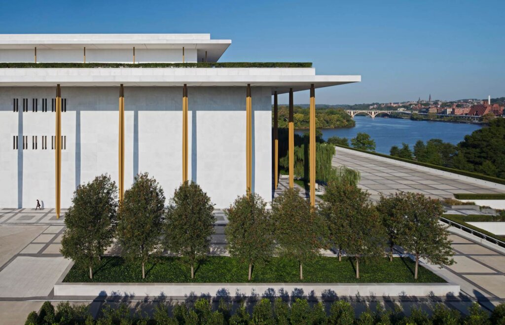 An exterior view of the John F. Kennedy Center for the Performing Arts in Washington, DC, a large, white building with a row of trees in front, on a sunny day.