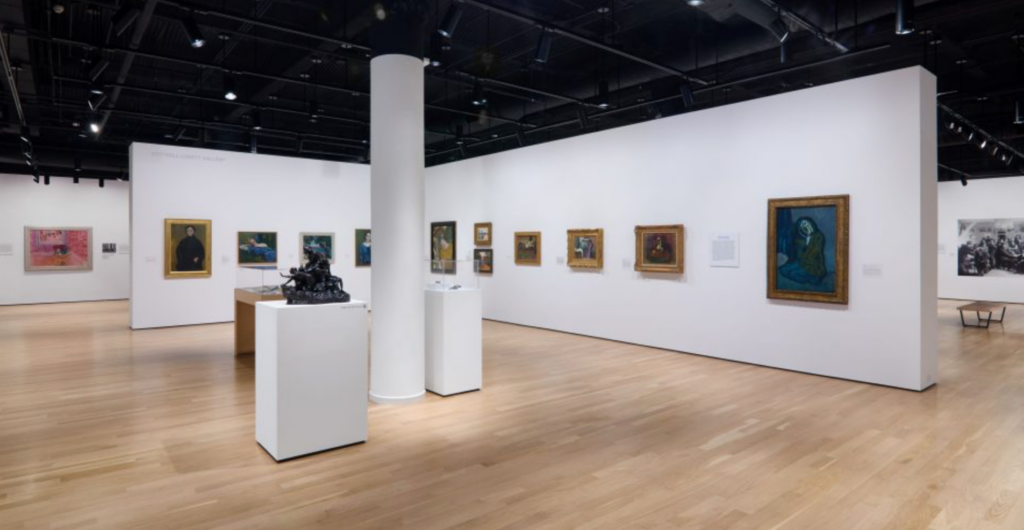 Installation view of 'Make Way for Berthe Weill' at NYU Grey Art Museum, an open gallery space with a black ceiling, white walls, and light wood floors. Paintings are hung on the wall, with a sculpture and smaller works in vitrines standing in the center of the gallery.