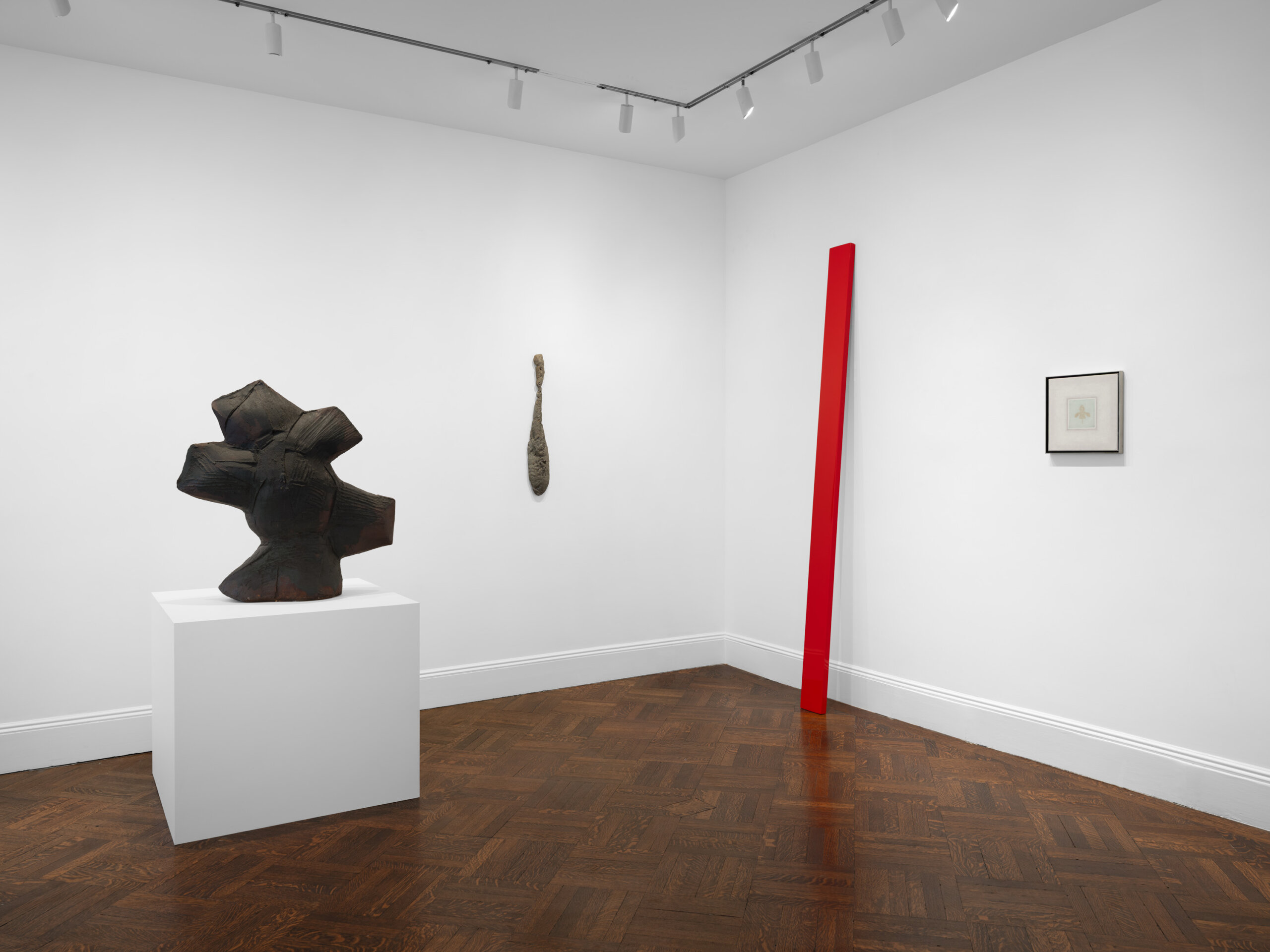 exhibition image. blank white walls with 4 art pieces. Far left is a black asymmetrical sculpture. Center is an object hanging from the ceiling and a tall red board/ sculpture leaning on the right wall. Then a small painting next to the red sculpture on the right side.