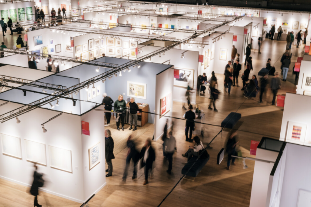 Image of park avenue armory at the IFPDA Print Fair in 2024. It shows stalls of art work with busy people walking all around. The image is from above.