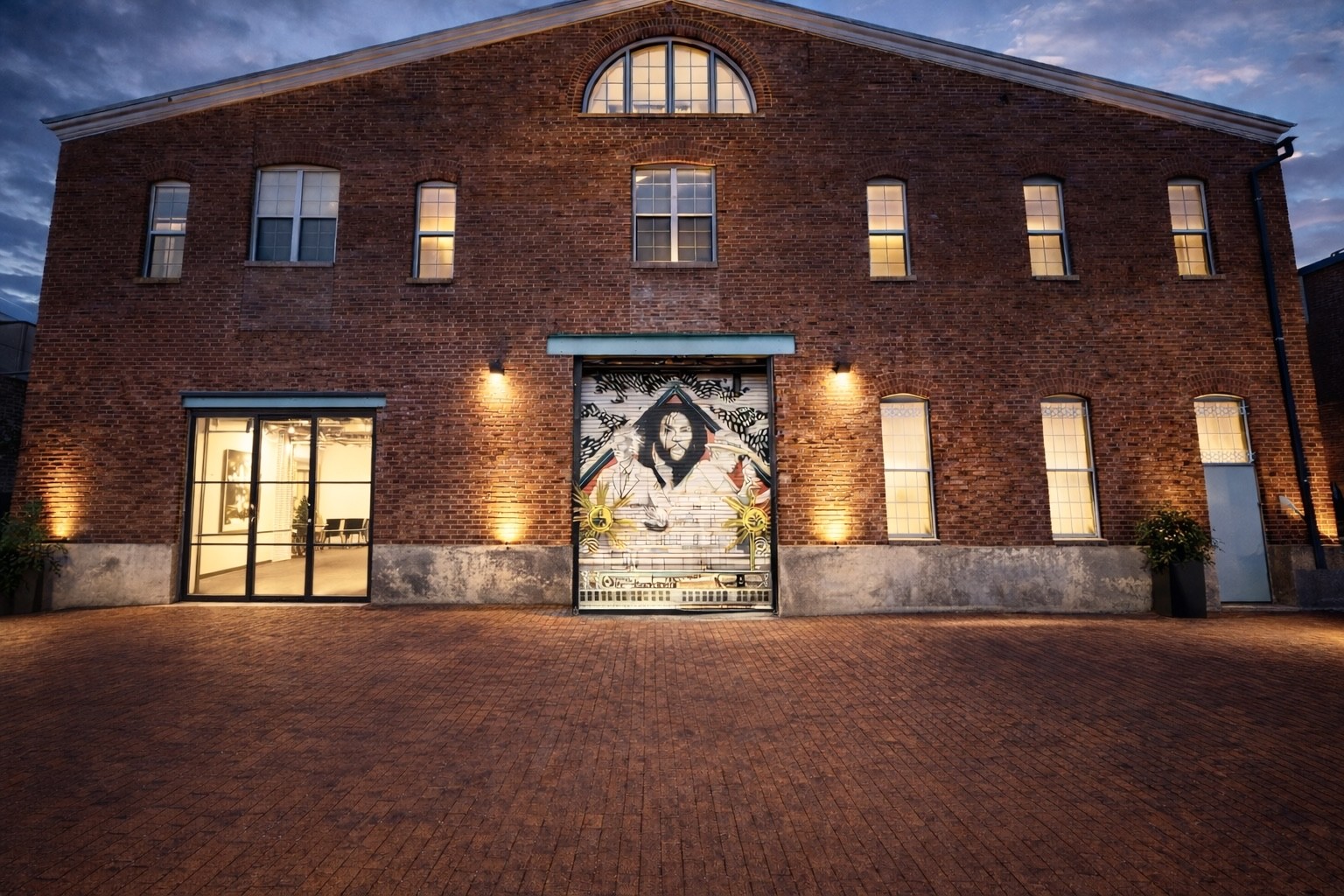 External shot of NCO Gallery. It is a brick building with many windows. 2 story with a larger window/door on the left side that opens to the gallery. There is also a freight door that has a mural on it.
